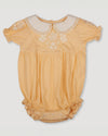 Early Sunday, Matilda Baby Bubble Romper in Vanilla color, made from Cotton Dobby Swiss Dots fabric, featuring ruffled neckline for a delicate touch and elasticated leg openings for a comfortable fit, shown in a front flat-lay view