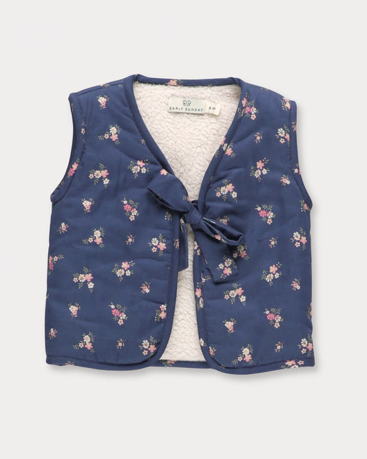 Early Sunday, Milly Baby Quilted Gilet in Ditsy Bouquet Navy, made from Printed Cotton Satin fabric, featuring a sleeveless jacket with a bow tie, shown in a front flat-lay view