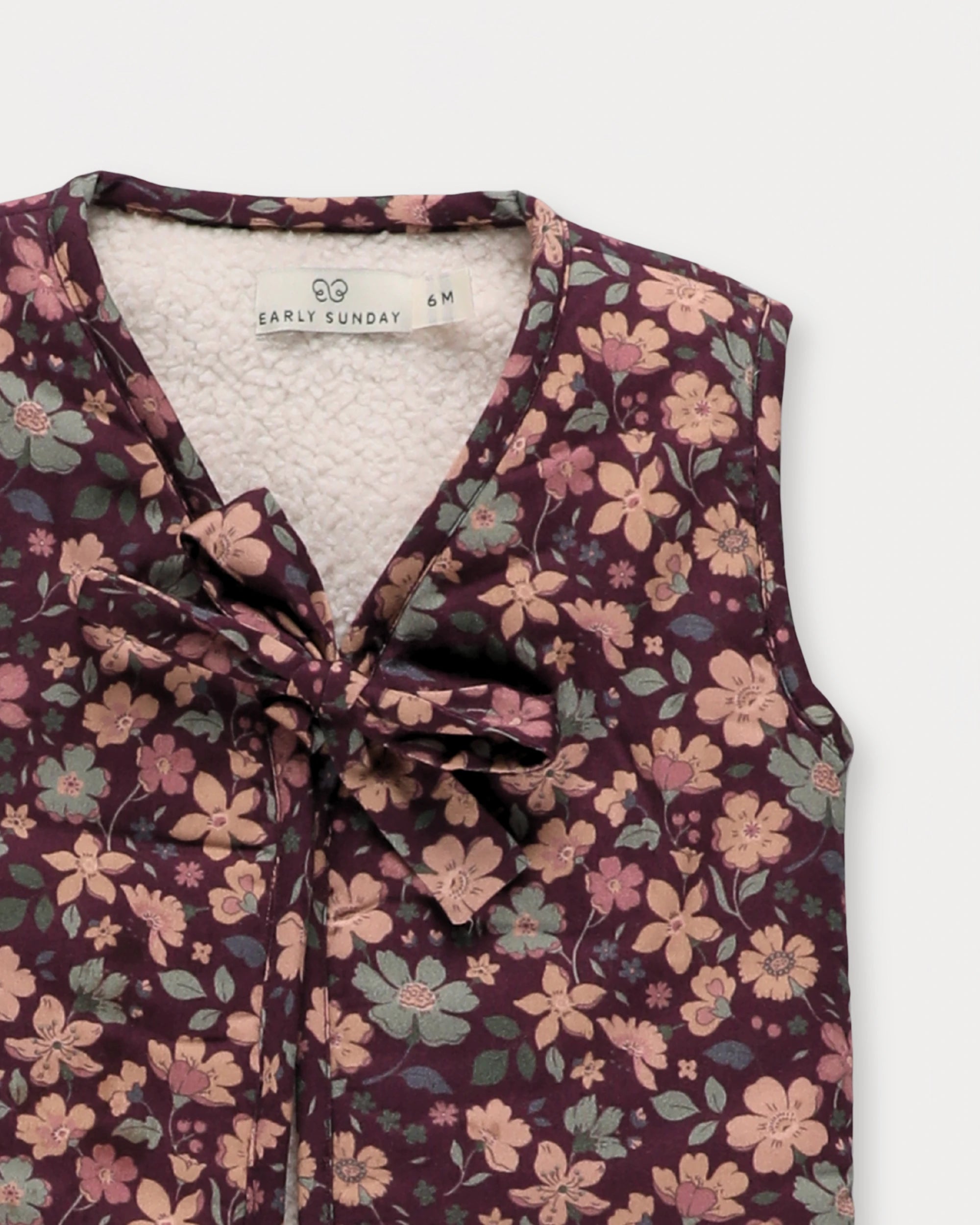 Early Sunday, Milly Baby Quilted Gilet in Garden Flowers Plum, made from Printed Cotton Satin fabric, featuring a sleeveless jacket with a bow tie, shown in a closeup flat-lay view