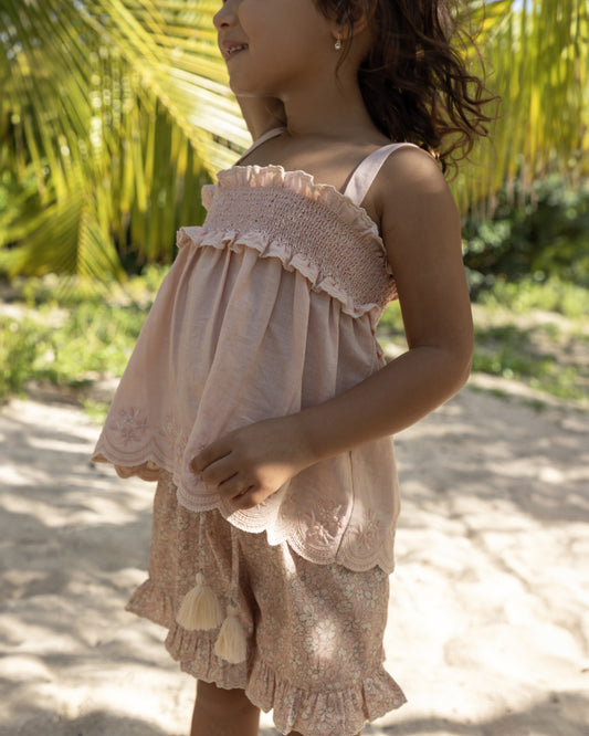 Early Sunday, Olivia Blouse in Pink Sand color, made from Canvas 70% Cotton 30% Linen fabric, featuring smocked bodice for a flexible fit, cross-back strap sand a flared hem, shown in a side view of model wearing it