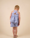Early Sunday, Romy Blouse in Summer Sky color, made from  100% Cotton Chambray fabric, featuring a sleeveless design with cross-back straps, a gathered waist & a beautiful embroidery, shown in a back view of a model wearing it in a studio setting