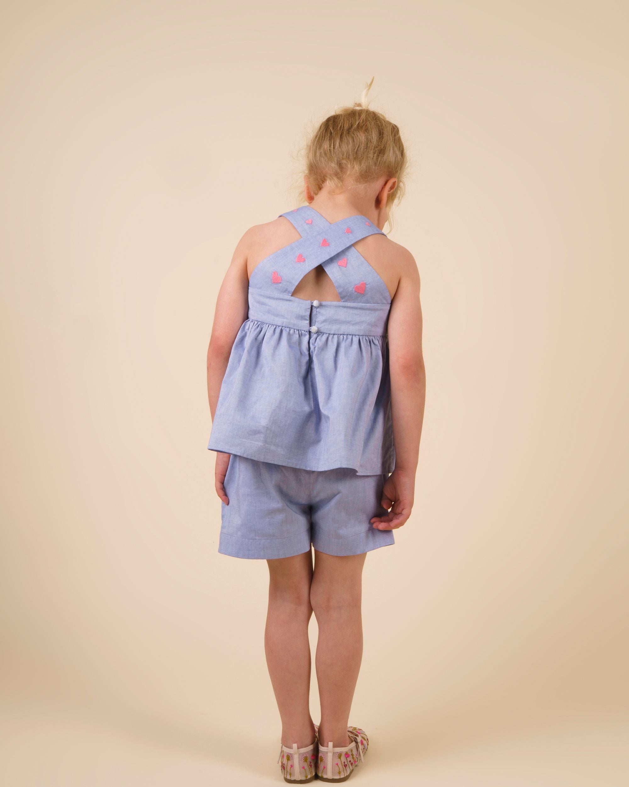 Early Sunday, Romy Blouse in Summer Sky color, made from  100% Cotton Chambray fabric, featuring a sleeveless design with cross-back straps, a gathered waist & a beautiful embroidery, shown in a back view of a model wearing it in a studio setting