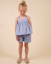Early Sunday, Romy Blouse in Summer Sky color, made from  100% Cotton Chambray fabric, featuring a sleeveless design with cross-back straps, a gathered waist & a beautiful embroidery, shown in a front view of a model wearing it in a studio setting