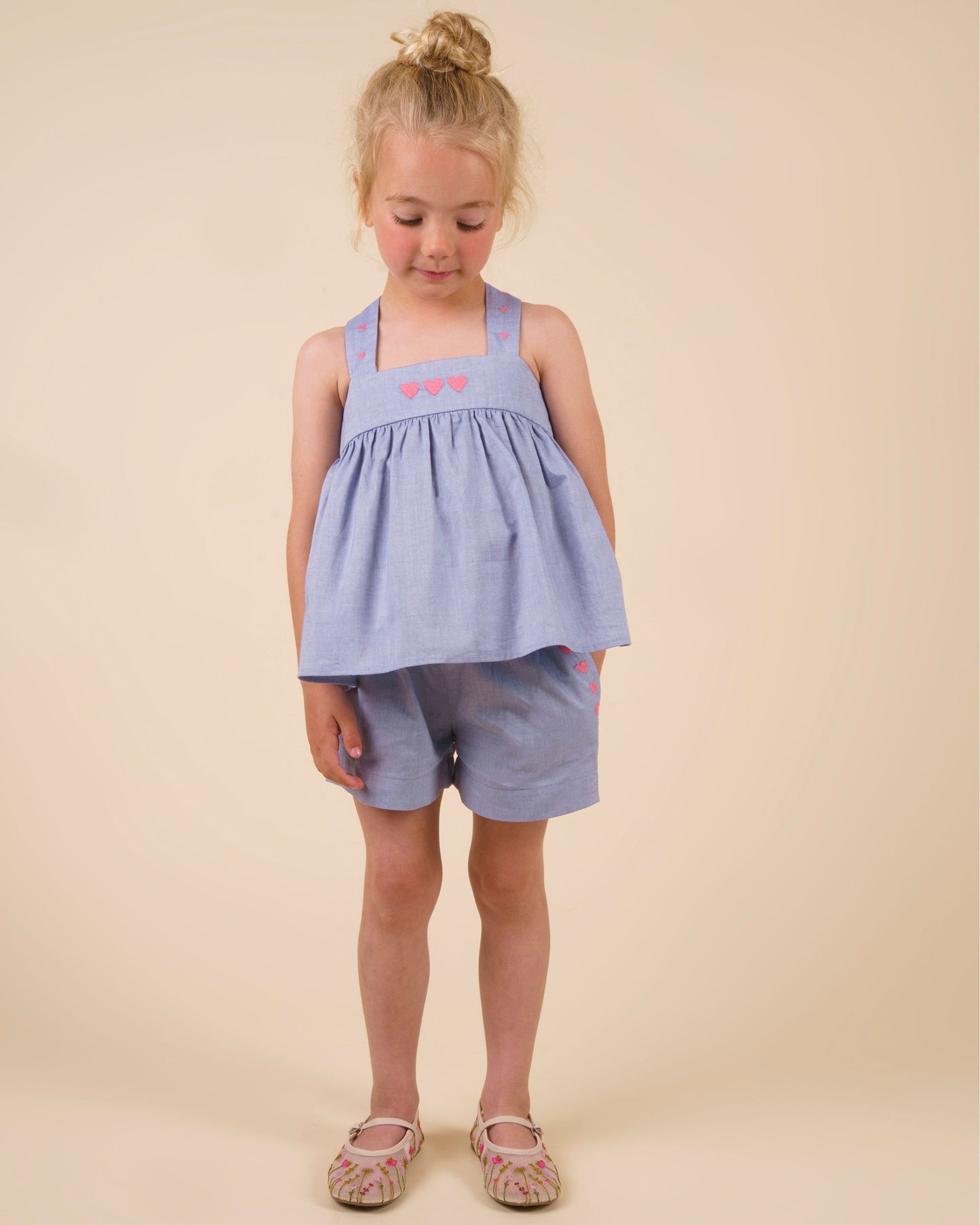 Early Sunday, Romy Blouse in Summer Sky color, made from  100% Cotton Chambray fabric, featuring a sleeveless design with cross-back straps, a gathered waist & a beautiful embroidery, shown in a front view of a model wearing it in a studio setting