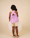 Early Sunday, Romy Blouse in Lilac color, made from 70% Cotton 30% Linen fabric, featuring a sleeveless design with cross-back straps, a gathered waist & a beautiful embroidery, shown in a back view of a model wearing it in a studio setting