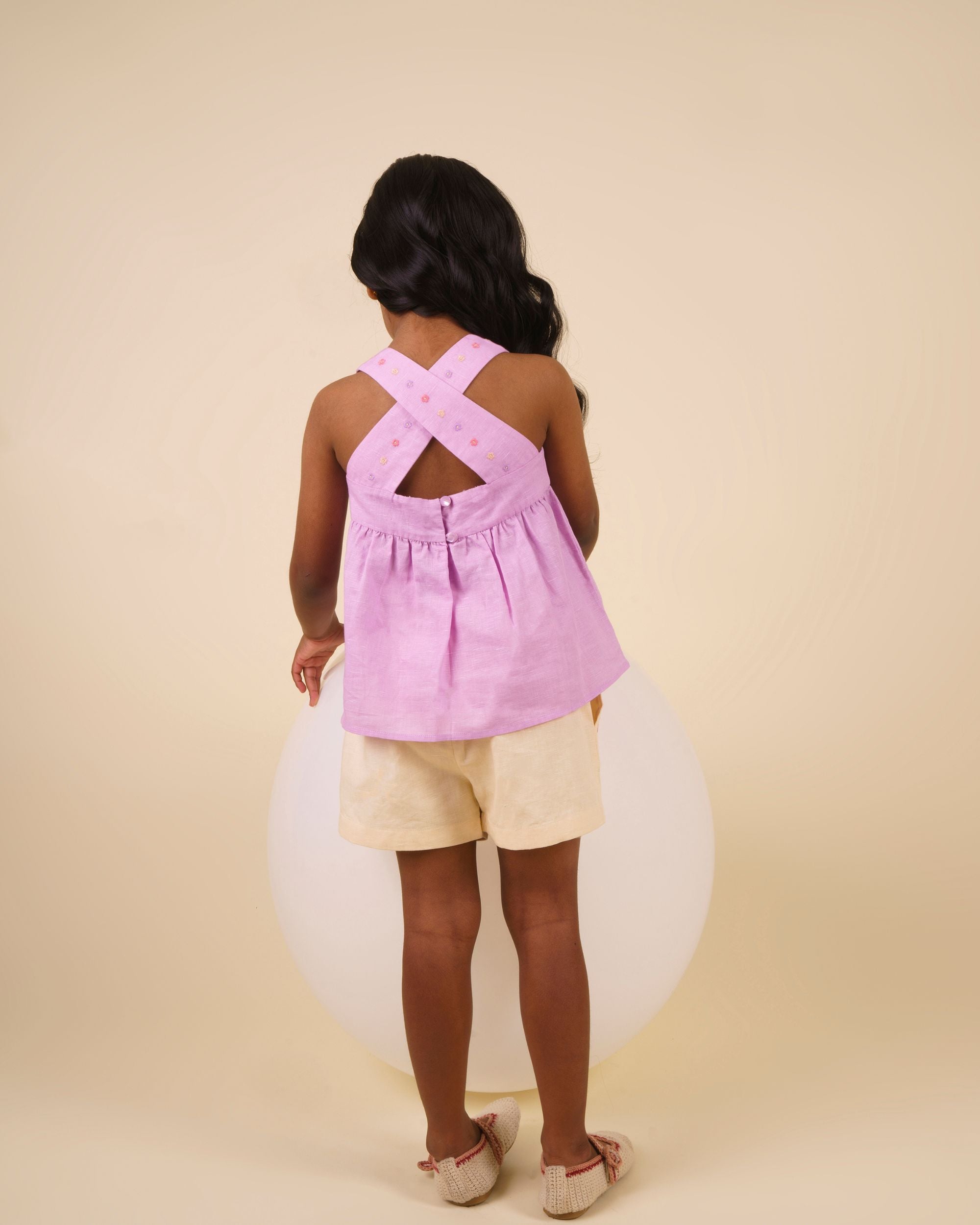 Early Sunday, Romy Blouse in Lilac color, made from 70% Cotton 30% Linen fabric, featuring a sleeveless design with cross-back straps, a gathered waist & a beautiful embroidery, shown in a back view of a model wearing it in a studio setting