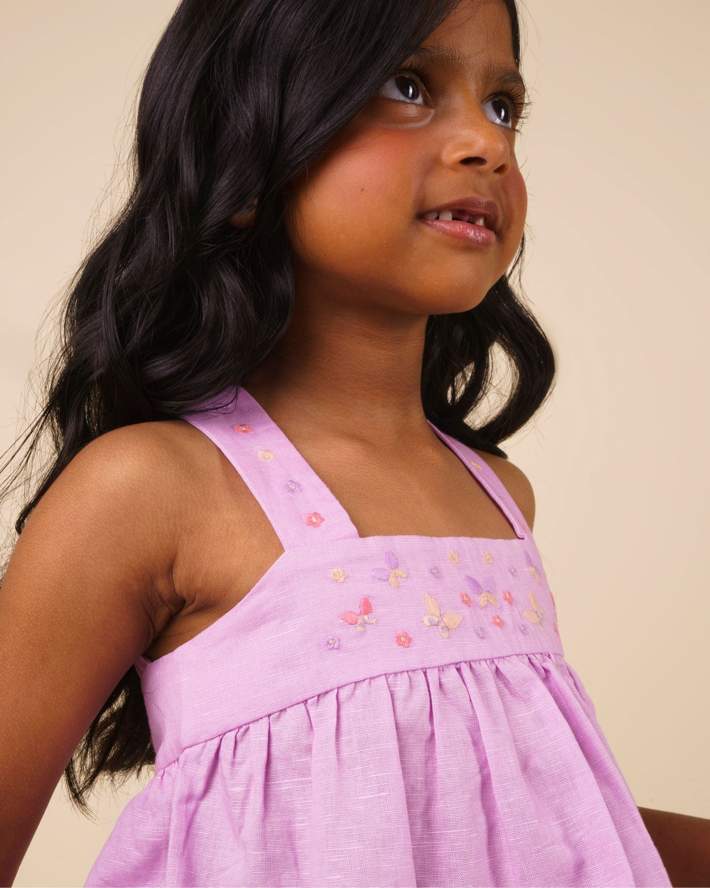 Early Sunday, Romy Blouse in Lilac color, made from 70% Cotton 30% Linen fabric, featuring a sleeveless design with cross-back straps, a gathered waist & a beautiful embroidery, shown in a close-up shot of a model wearing it during a studio shoot