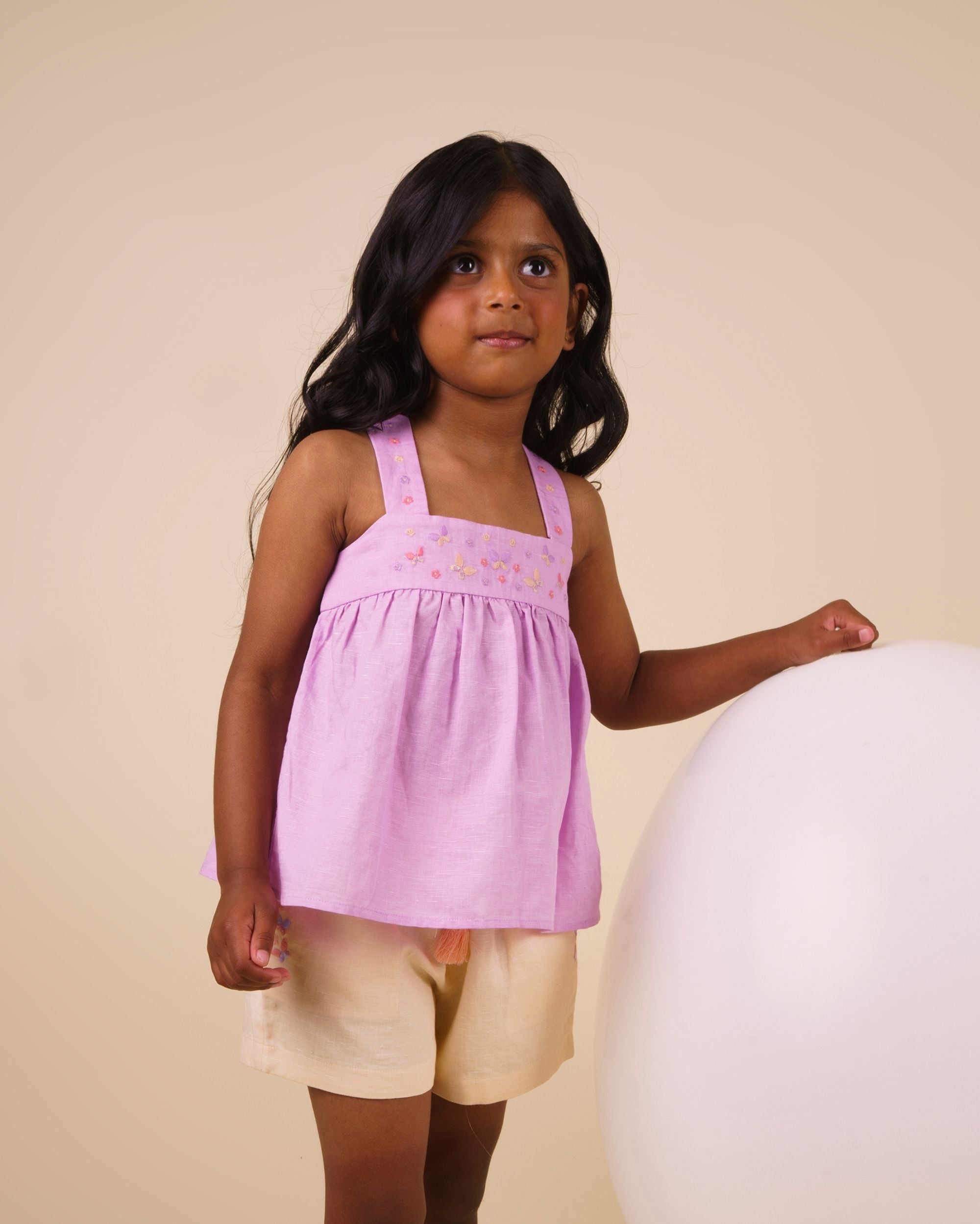 Early Sunday, Romy Blouse in Lilac color, made from 70% Cotton 30% Linen fabric, featuring a sleeveless design with cross-back straps, a gathered waist & a beautiful embroidery, shown in a front view of a model wearing it in a studio setting