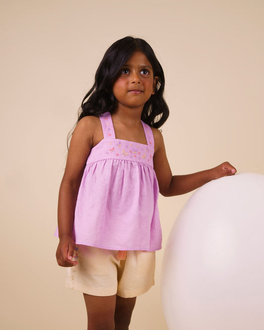 Early Sunday, Romy Blouse in Lilac color, made from 70% Cotton 30% Linen fabric, featuring a sleeveless design with cross-back straps, a gathered waist & a beautiful embroidery, shown in a front view of a model wearing it in a studio setting
