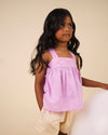 Early Sunday, Romy Blouse in Lilac color, made from 70% Cotton 30% Linen fabric, featuring a sleeveless design with cross-back straps, a gathered waist & a beautiful embroidery, shown in an angled view of a model wearing it in a studio setting