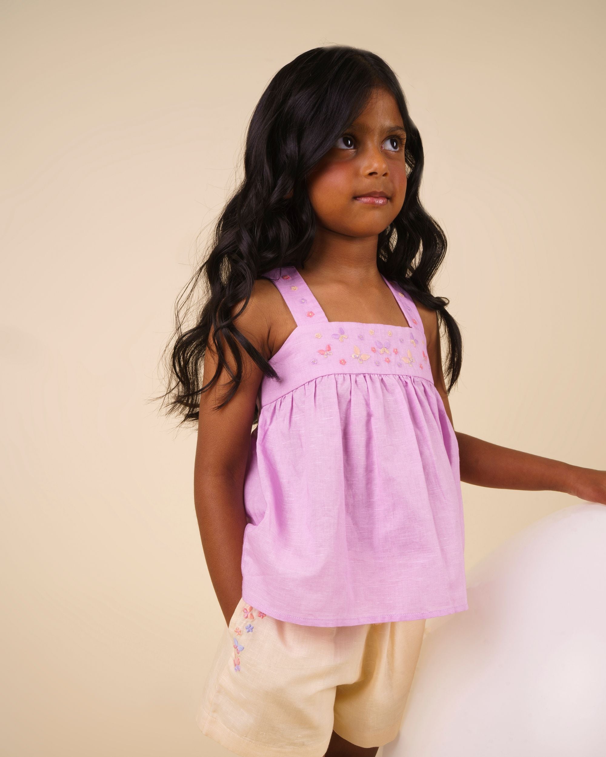 Early Sunday, Romy Blouse in Lilac color, made from 70% Cotton 30% Linen fabric, featuring a sleeveless design with cross-back straps, a gathered waist & a beautiful embroidery, shown in an angled view of a model wearing it in a studio setting