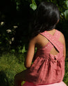 Early Sunday, Romy Blouse in Peony color, made from 70% Cotton 30% Linen fabric, featuring a sleeveless design with cross-back straps, a gathered waist & a beautiful embroidery, shown in a back view of a model wearing it in a studio setting