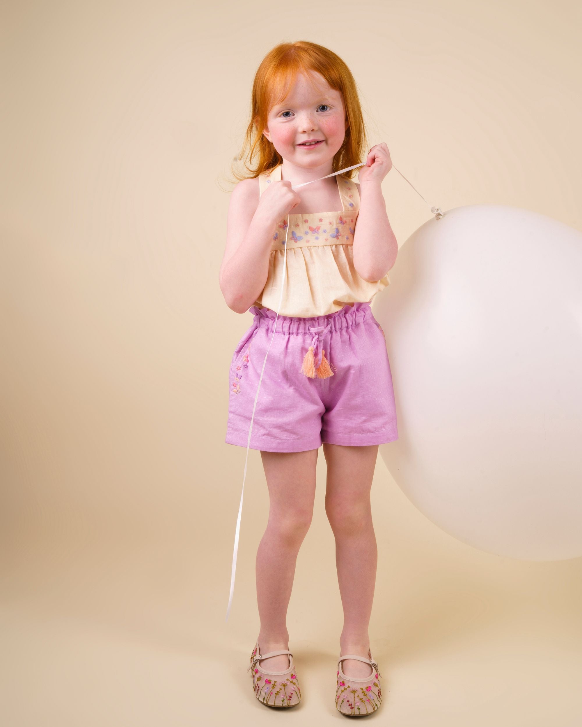 Early Sunday, Romy Blouse in Sand color, made from 70% Cotton 30% Linen fabric, featuring a sleeveless design with cross-back straps, a gathered waist & a beautiful embroidery, shown in a front view of a model wearing it in a studio setting