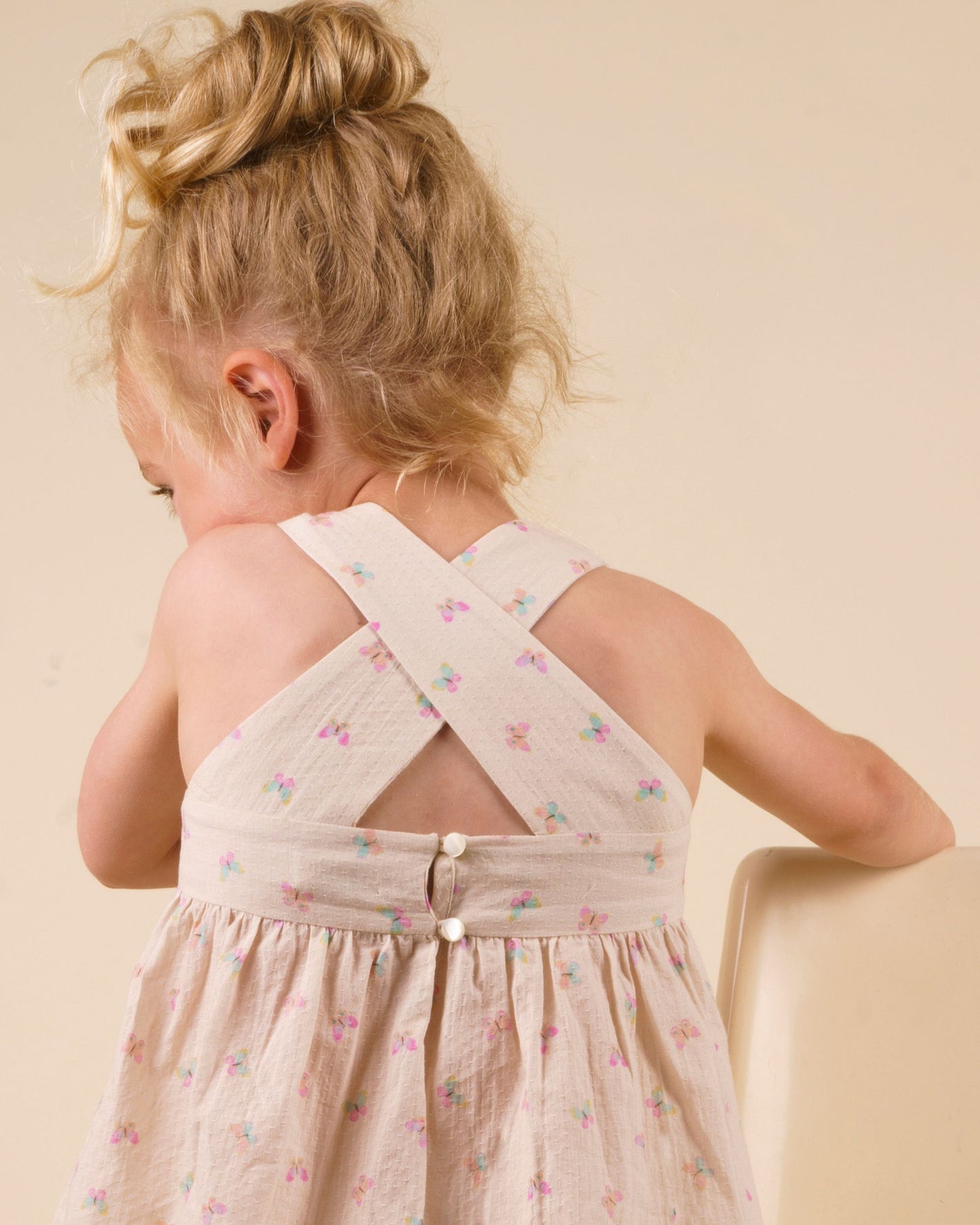 Early Sunday, Romy Blouse in Butterflies print, made from Printed Cotton Tiny Swiss Dobby fabric, featuring a sleeveless design with cross-back straps, a gathered waist & a soft flared silhouette, shown in close-up back view of a model wearing it in a studio setting