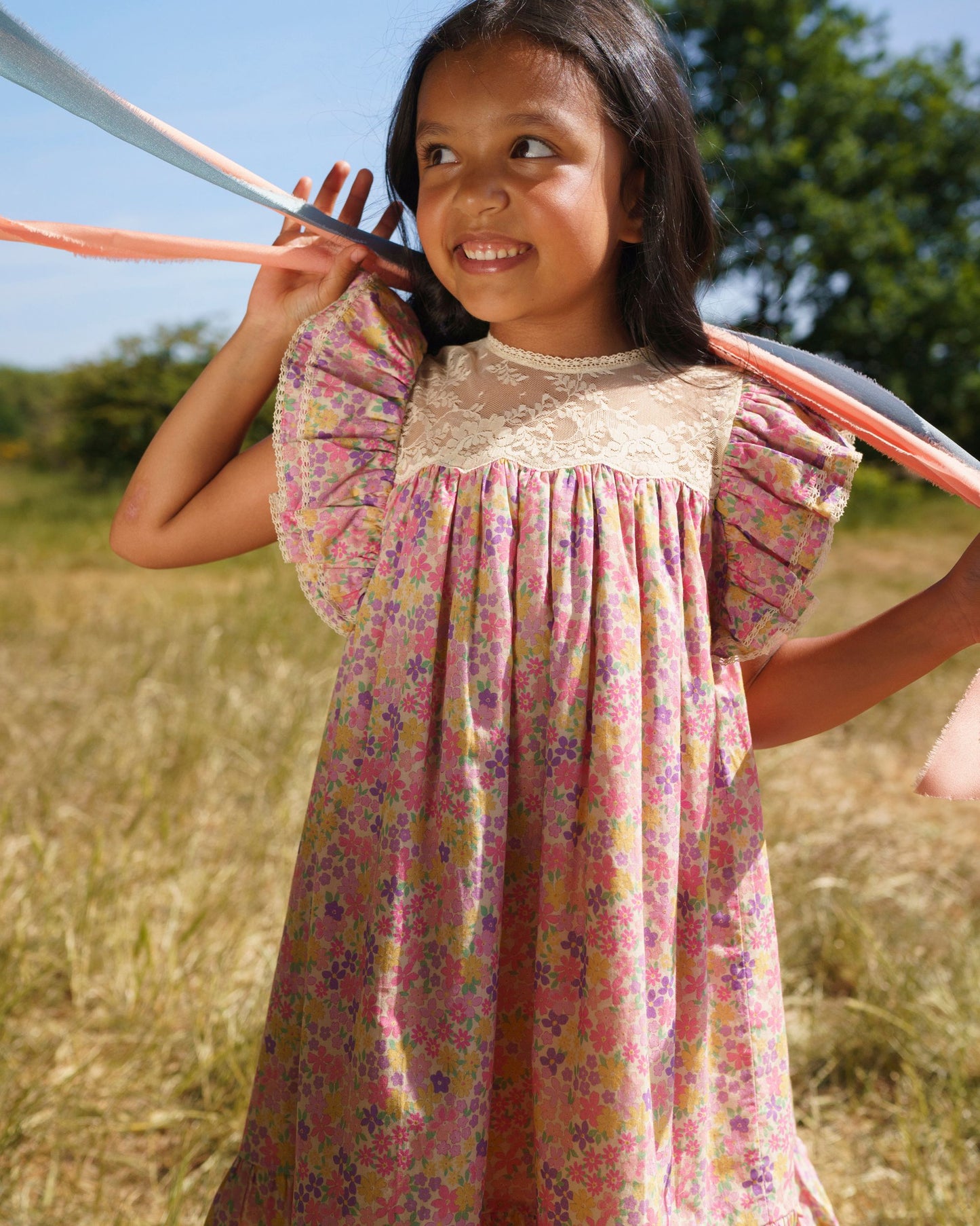 Early Sunday, Sita Dress in Emma Floral Purple color, made from Organic Printed Cotton Cambric fabric, featuring a sleeveless design with wide shoulder straps & a delicate lace yoke, shown in a close up shot of model playing outdoors