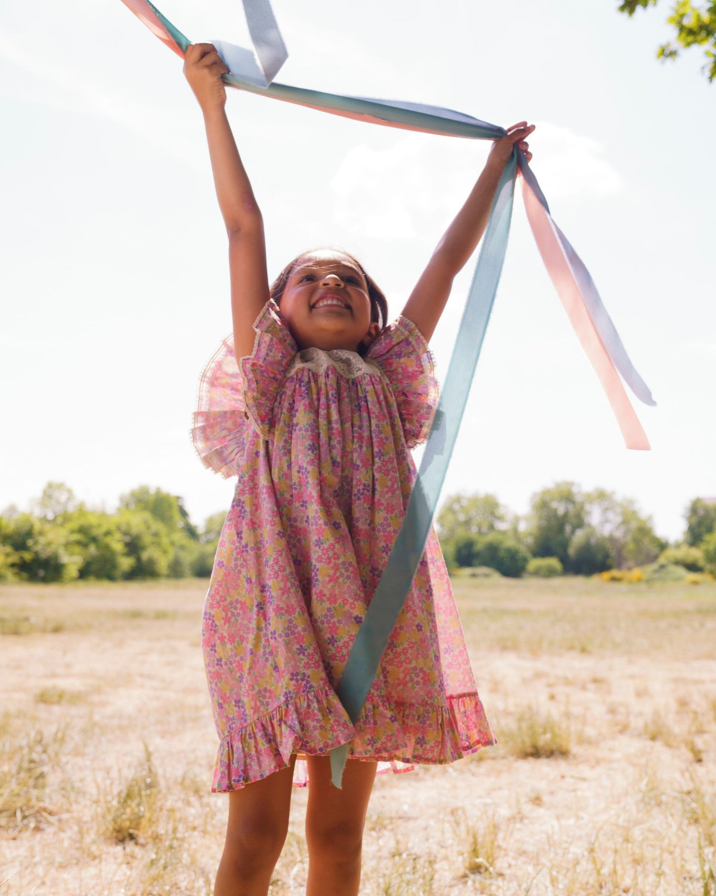 Early Sunday, Sita Dress in Emma Floral Purple color, made from Organic Printed Cotton Cambric fabric, featuring a sleeveless design with wide shoulder straps & a delicate lace yoke, shown in a front view of model playing outdoors