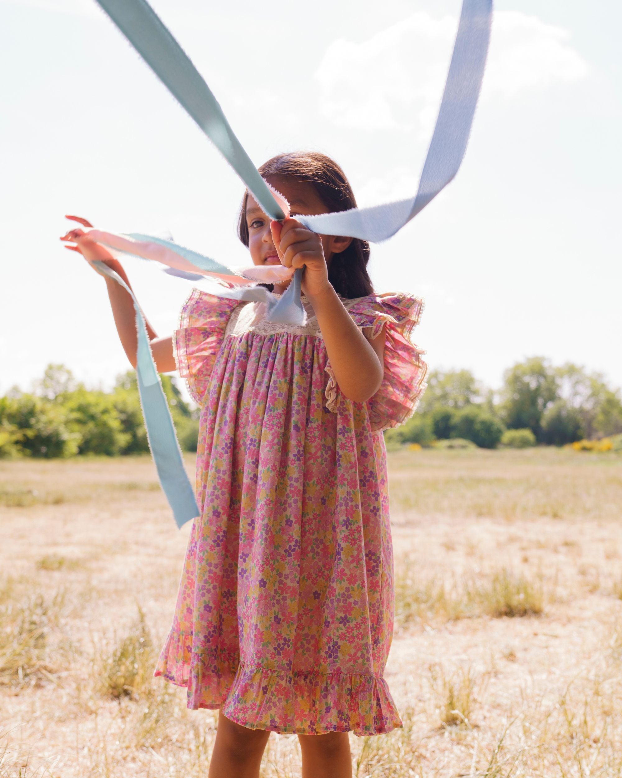 Early Sunday, Sita Dress in Emma Floral Purple color, made from Organic Printed Cotton Cambric fabric, featuring a sleeveless design with wide shoulder straps & a delicate lace yoke, shown in an angled view of a model wearing outdoors