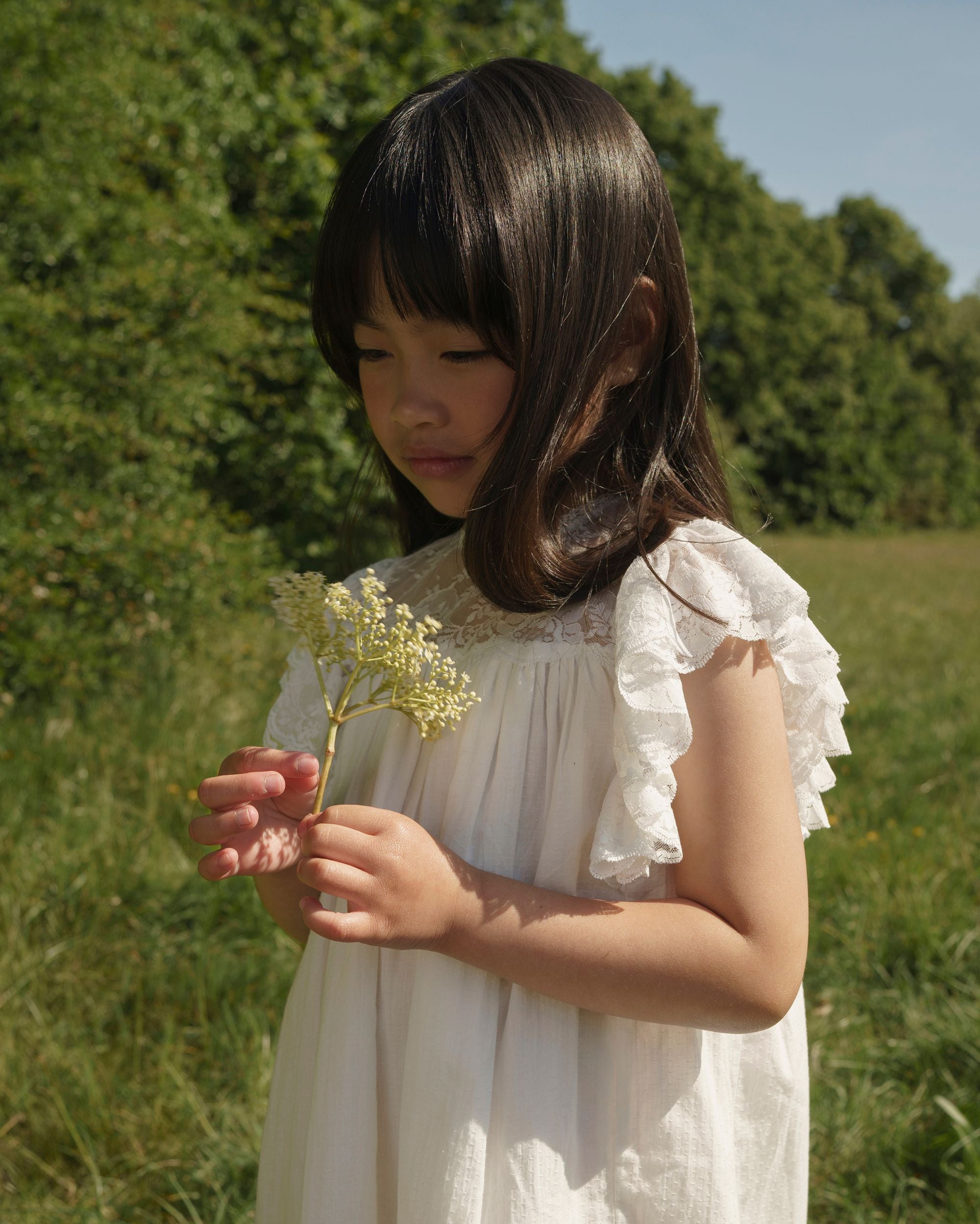 Early Sunday, Sita Dress in Ivory color, made from Tiny Swiss Dobby fabric, featuring a sleeveless design with wide shoulder straps & a delicate lace yoke, shown in a close up shot of model playing outdoors