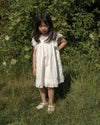 Early Sunday, Sita Dress in Ivory color, made from Tiny Swiss Dobby fabric, featuring a sleeveless design with wide shoulder straps & a delicate lace yoke, shown in a front view of model wearing outdoors