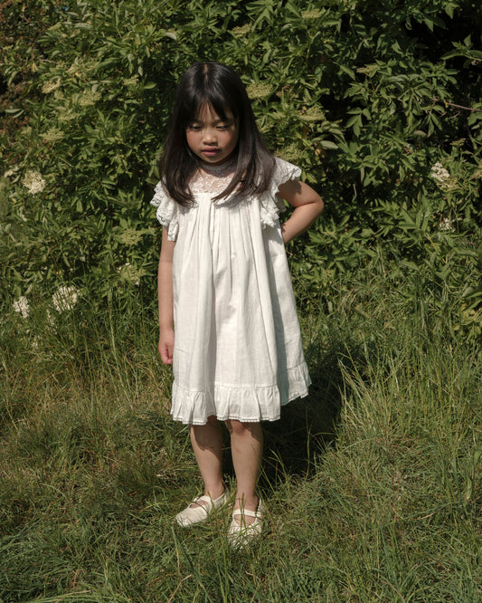 Early Sunday, Sita Dress in Ivory color, made from Tiny Swiss Dobby fabric, featuring a sleeveless design with wide shoulder straps & a delicate lace yoke, shown in a front view of model wearing outdoors
