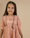Early Sunday, Suzie Dress in Acorn Dusty Pink, made from Printed Cotton Double Gauze fabric, featuring classic silhouette with a ruffle neckline and mother-of-pearl buttons, shown in a close-up shot of a model wearing it during a studio shoot